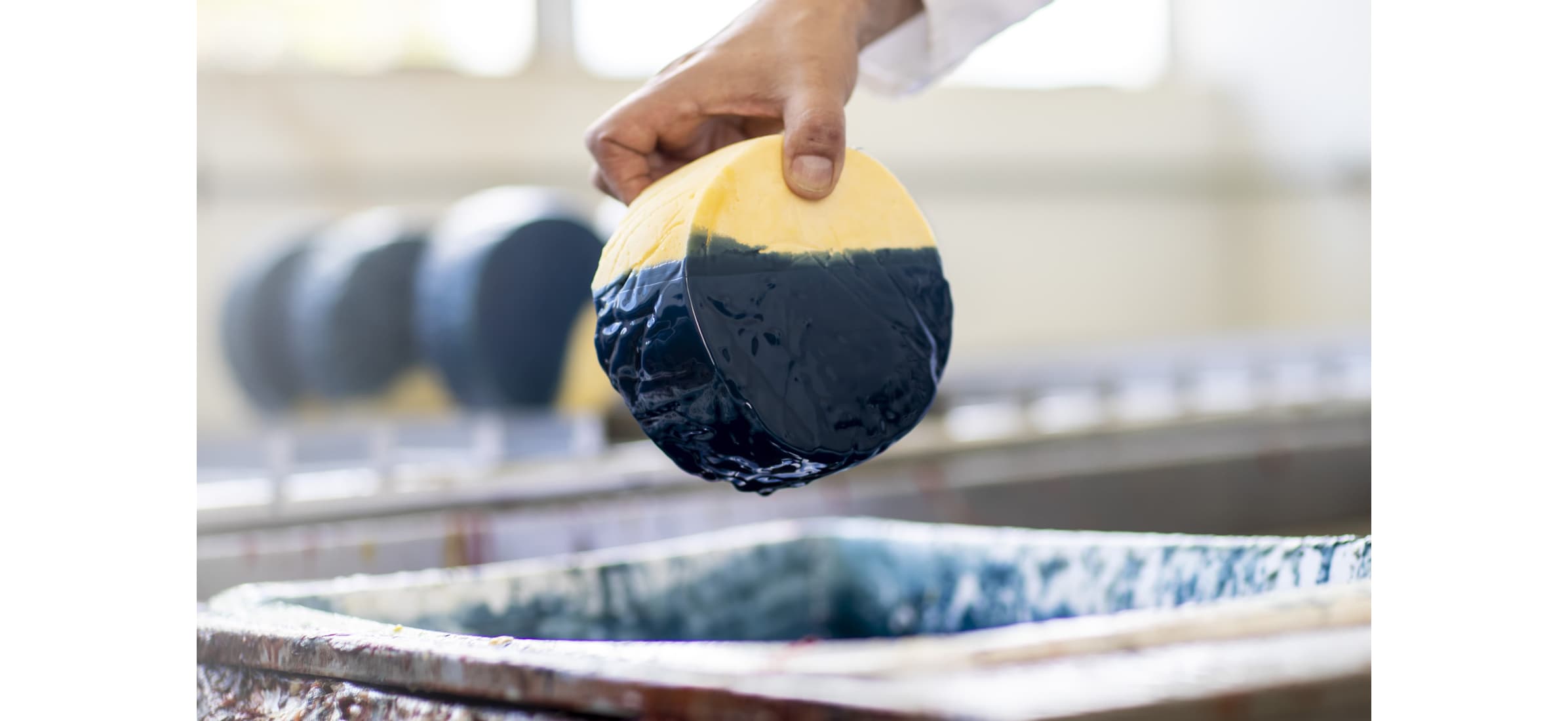 A hand dips a circular cheese block into blue wax.